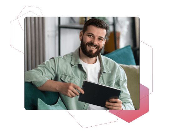 Ein bärtiger Mann im lässigen Outfit sitzt mit einem Tablet auf einem Sofa und lächelt in die Kamera